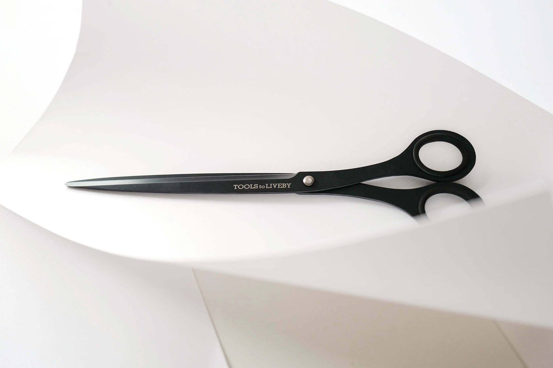 TOOLS to LIVEBY matte black scissors on a minimalist curved white paper background.