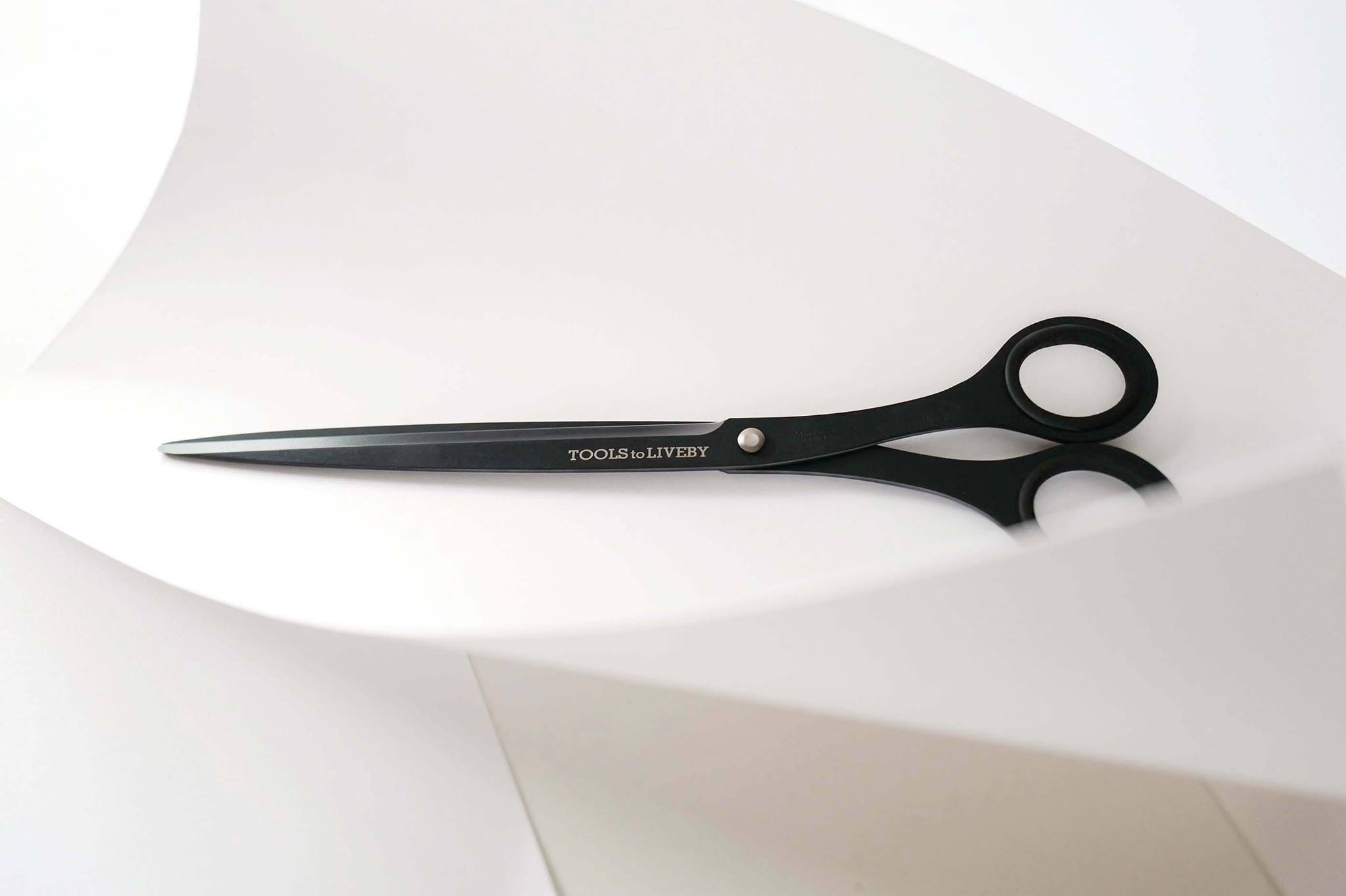 TOOLS to LIVEBY matte black scissors on a minimalist curved white paper background.