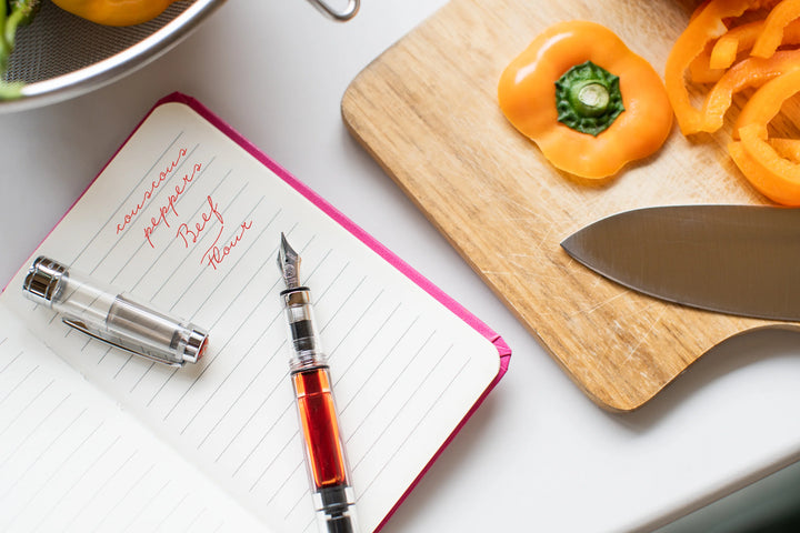 Notebook with TWSBI Diamond 580 Fountain Pen – Clear with filled red ink, sliced bell peppers on a cutting board, and knife on a white surface