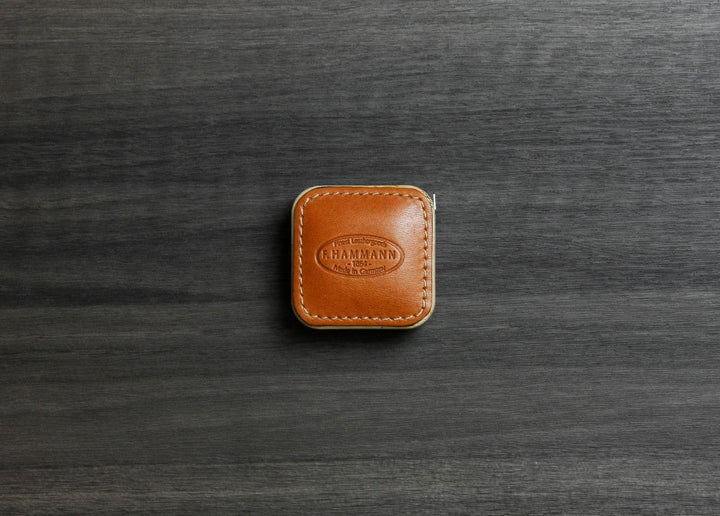 Top-down photograph of the F. Hammann 5205 Coxorange Alpina Leather Tape Measure resting on dark wood grain.