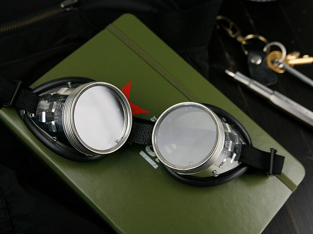 German Retro Safety Goggles with 50mm shatterproof lenses resting on an olive green notebook in a professional workshop setting.