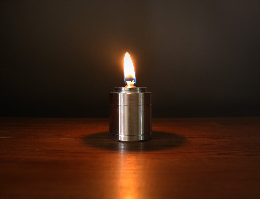 F Plus Duralumin Oil Candle — lit, casting a warm steady flame on a dark wooden surface