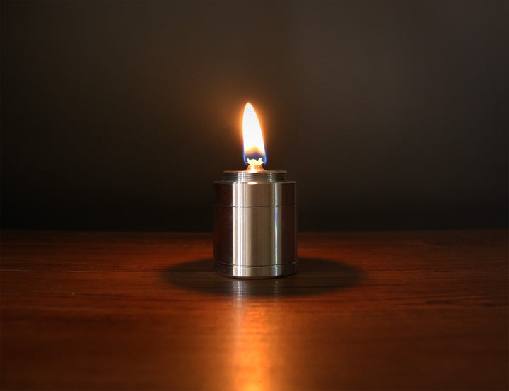 F Plus Duralumin Oil Candle — lit, casting a warm steady flame on a dark wooden surface