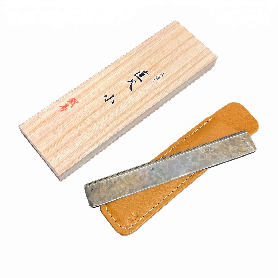 Compact 140mm Kurogane Kazura metal ruler next to its custom tan leather sleeve and traditional wooden box.