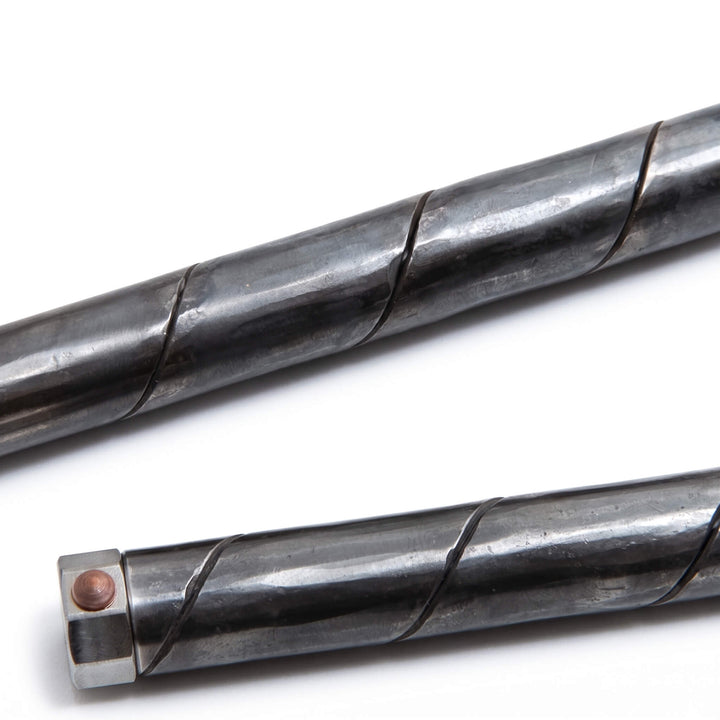 Macro detail of the raw fire-and-anvil forged finish and maki-hari rolled seam on a dark steel pen barrel.