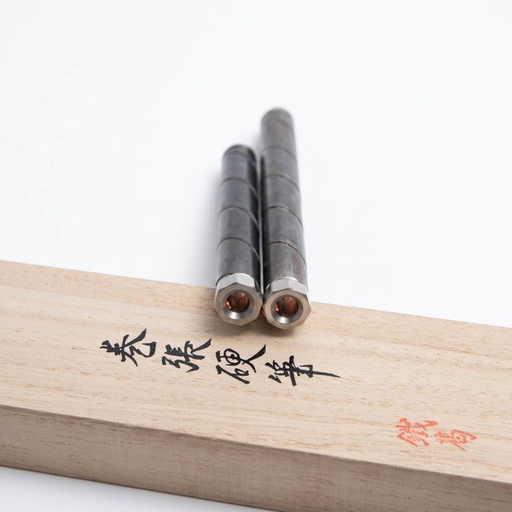 Two Japanese blacksmith-crafted everyday carry pens resting on a calligraphed gift box, showing hollow octagonal tail plugs.