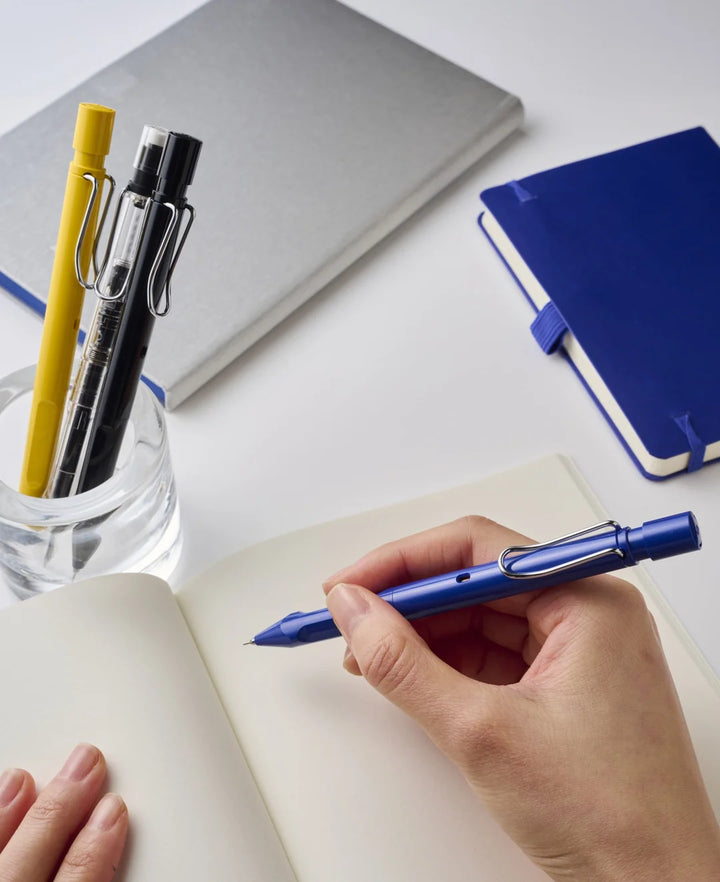 A person writing in a notebook using the blue LAMY safari KURUTOGA mechanical pencil, demonstrating the ergonomic grip, with the yellow and Vista Clear models resting in a glass cup on the desk.