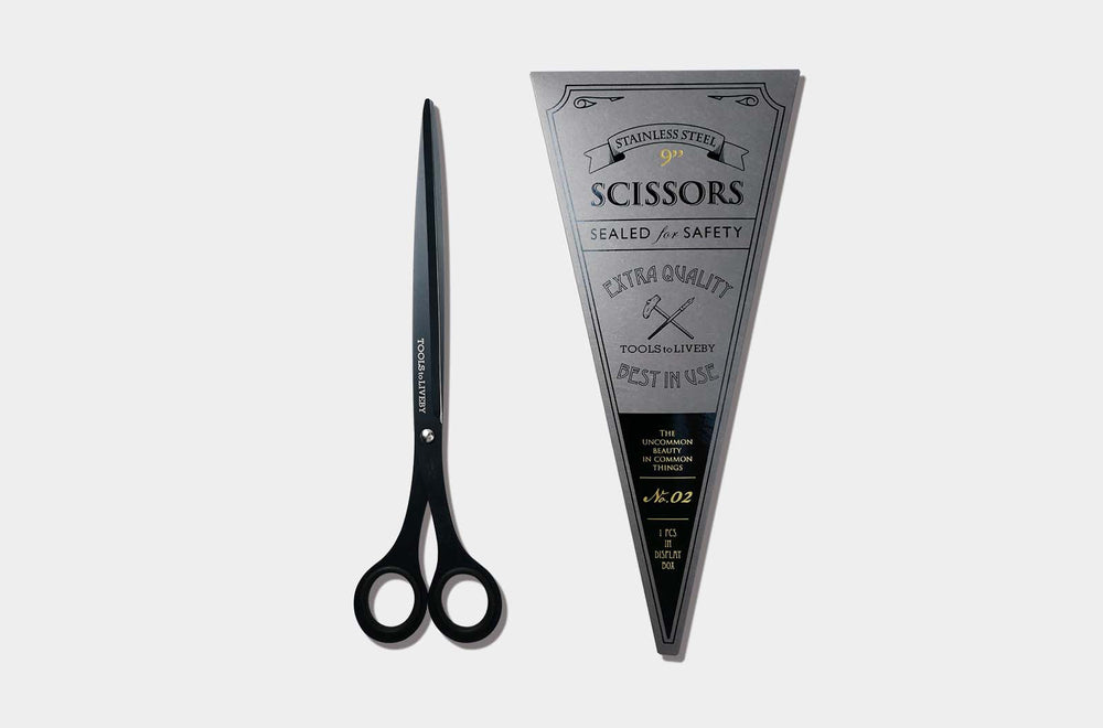 Overhead view of the elongated TOOLS to LIVEBY 9-inch matte black scissors placed vertically next to its premium No. 02 silver triangular cardboard packaging sleeve with vintage letterpress details.