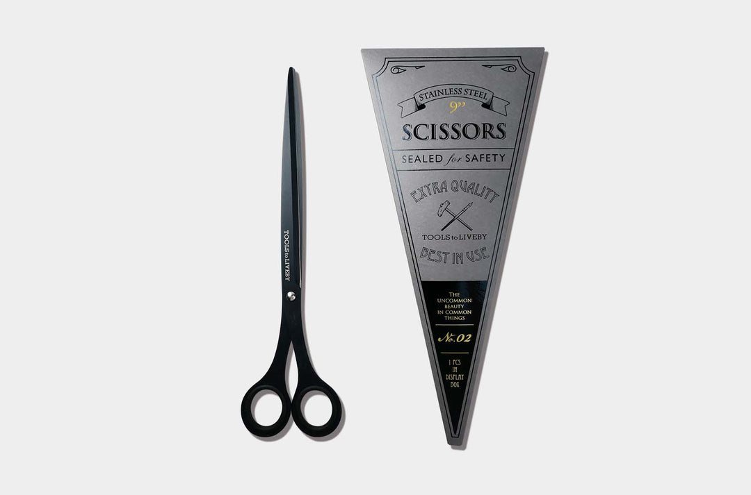 Overhead view of the elongated TOOLS to LIVEBY 9-inch matte black scissors placed vertically next to its premium No. 02 silver triangular cardboard packaging sleeve with vintage letterpress details.