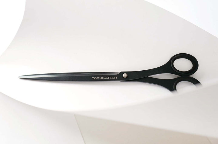 Close-up shot of the TOOLS to LIVEBY 9-inch matte black scissors effortlessly slicing through a thick sheet of curled white paper, demonstrating the razor-sharp Japanese steel blades and extended reach.
