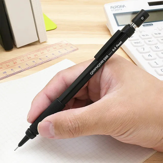 A hand holding the black Pentel GraphGear 500 PG515 mechanical pencil, positioned to write on graph paper with a ruler and calculator in the background.