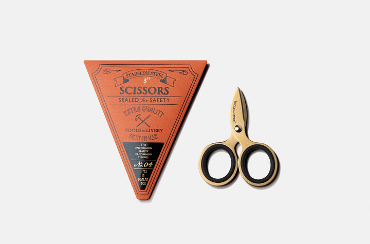 Overhead view of the luxurious TOOLS to LIVEBY 3-inch gold pocket scissors placed vertically next to its premium No. 04 terracotta triangular cardboard packaging sleeve with vintage letterpress details.