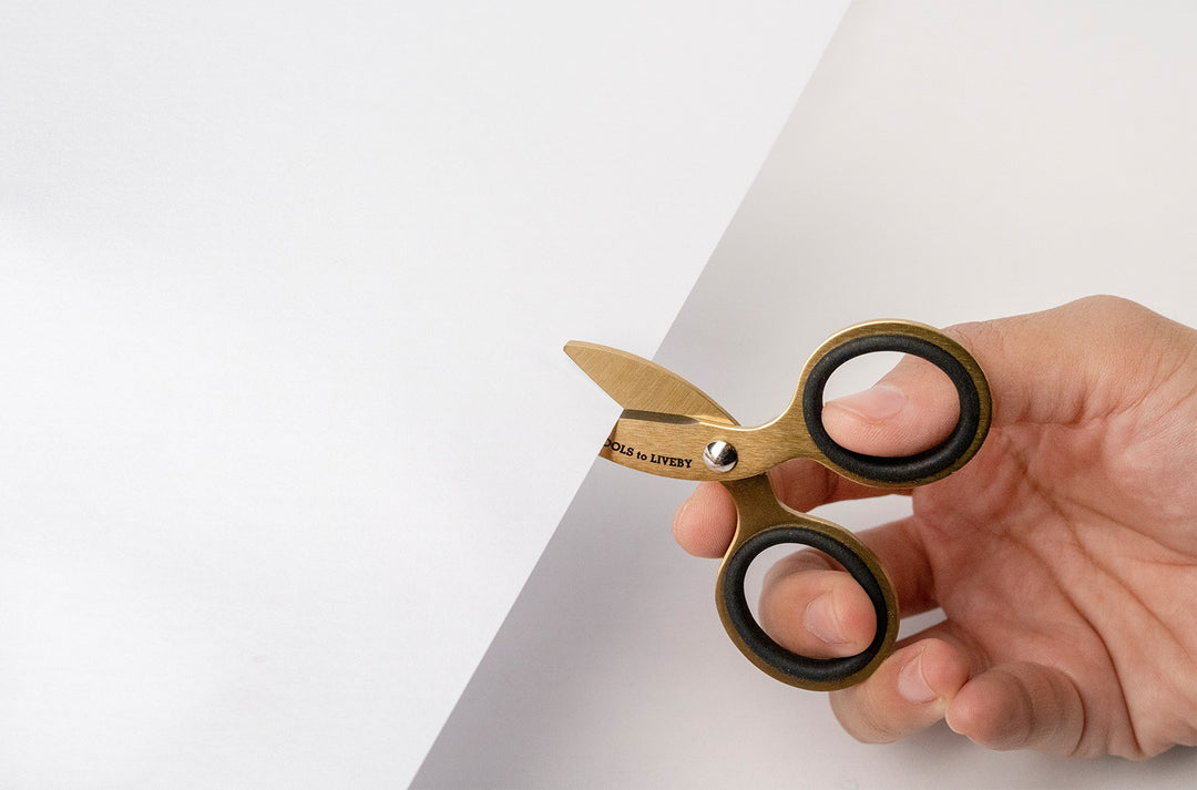 Close-up shot of a hand effortlessly making a precision cut into white paper using the ultra-compact TOOLS to LIVEBY 3-inch gold scissors, demonstrating the scale, ergonomic grip, and razor-sharp functionality of the luxurious micro-EDC tool.