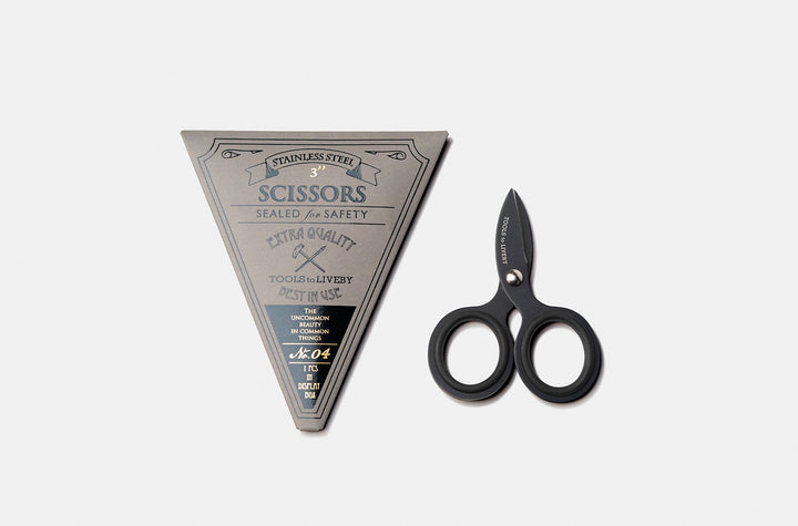 Overhead view of the ultra-compact TOOLS to LIVEBY 3-inch matte black pocket scissors placed vertically next to its premium No. 04 dark grey triangular cardboard packaging sleeve with vintage letterpress details.