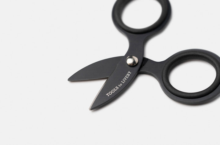 Macro close-up of the partially open TOOLS to LIVEBY 3-inch matte black scissors, displaying the sharp inner edge of the micro Japanese stainless steel blades, the silver tension screw, and the precise anti-adhesive Teflon coating.