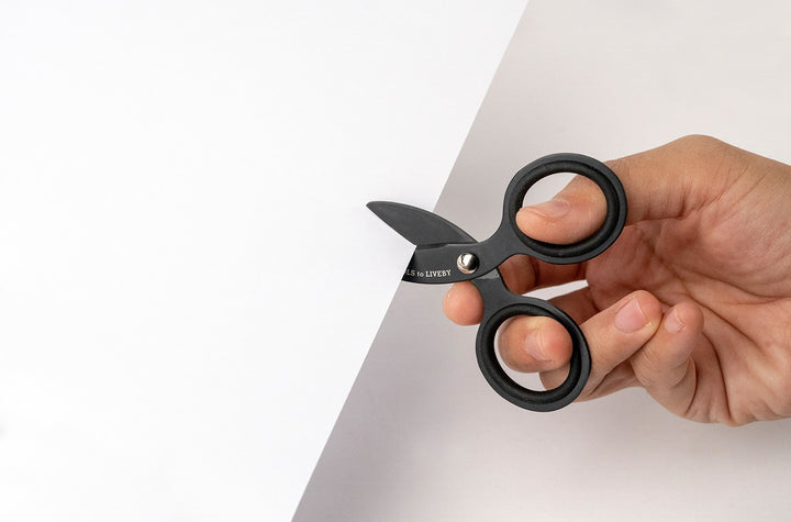 Close-up shot of a hand effortlessly making a precision cut into white paper using the ultra-compact TOOLS to LIVEBY 3-inch matte black scissors, demonstrating the scale, ergonomic grip, and razor-sharp functionality of the micro-EDC tool.