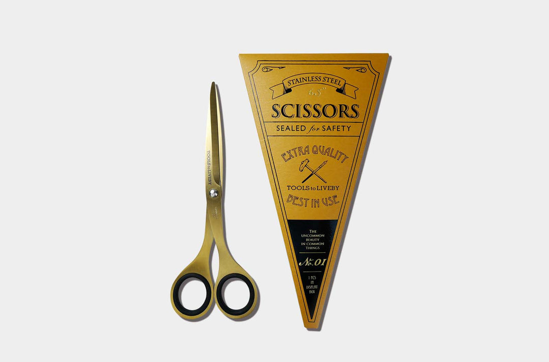 Overhead view of the compact TOOLS to LIVEBY 6.5-inch gold scissors placed vertically next to its premium No. 01 vibrant yellow triangular cardboard packaging sleeve with vintage letterpress details.