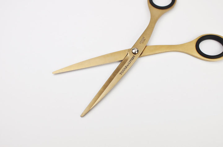 Macro close-up of the open TOOLS to LIVEBY 6.5-inch gold scissors, displaying the sharp inner edge of the Japanese stainless steel blades, the silver tension screw, and the brilliant golden finish.