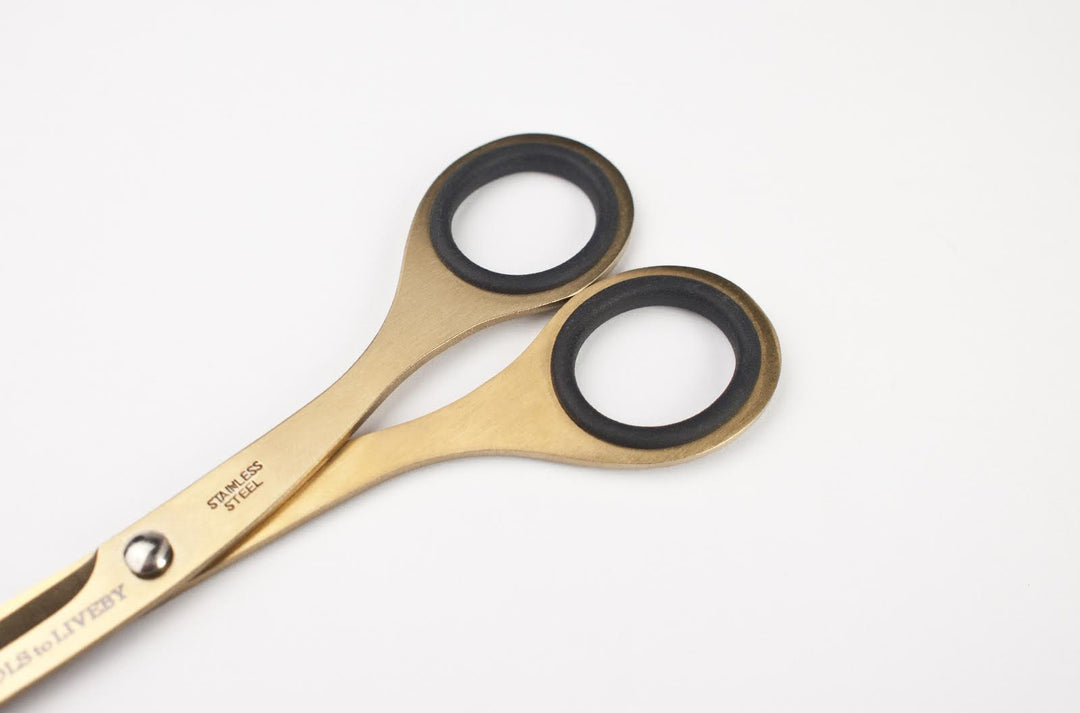 Macro detail of the TOOLS to LIVEBY 6.5-inch gold scissors' handles, highlighting the ergonomic black rubber inner rings, the engraved 'Stainless Steel' mark, and the eye-catching golden body.