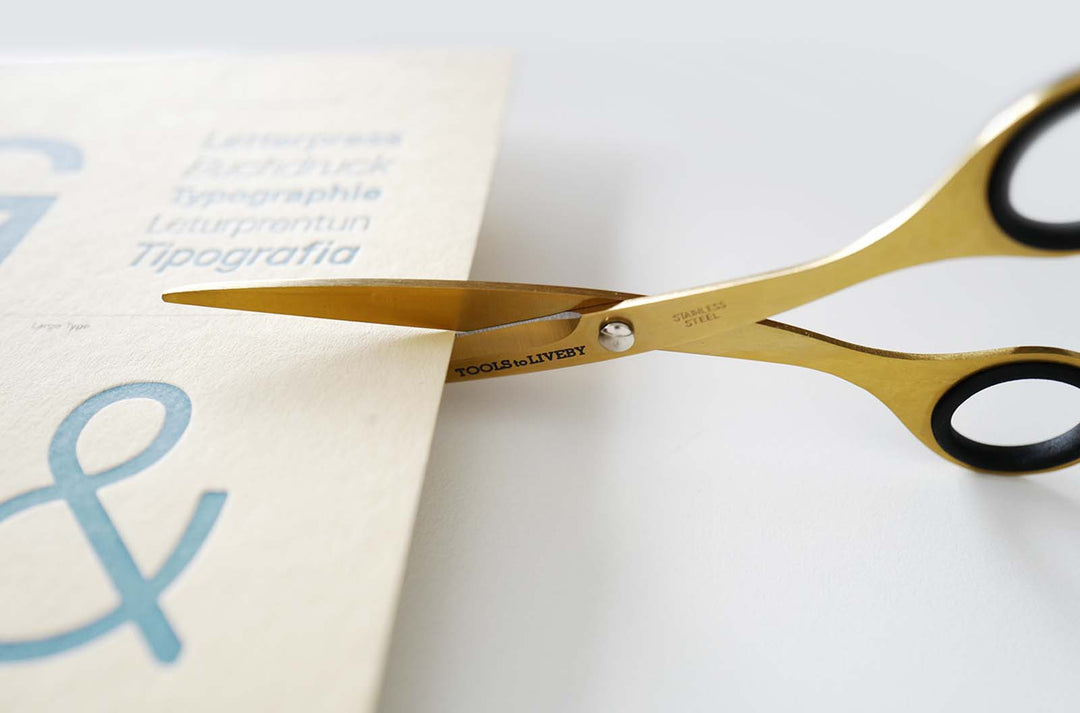 Close-up shot of the TOOLS to LIVEBY 6.5-inch gold scissors effortlessly slicing through a sheet of thick letterpress paper, demonstrating the razor-sharp Japanese steel blades in action.