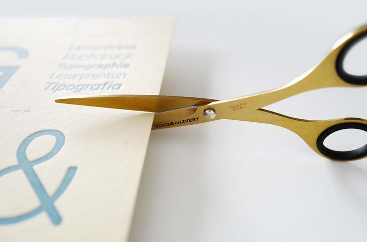 Close-up shot of the TOOLS to LIVEBY 6.5-inch gold scissors effortlessly slicing through a sheet of thick letterpress paper, demonstrating the razor-sharp Japanese steel blades in action.
