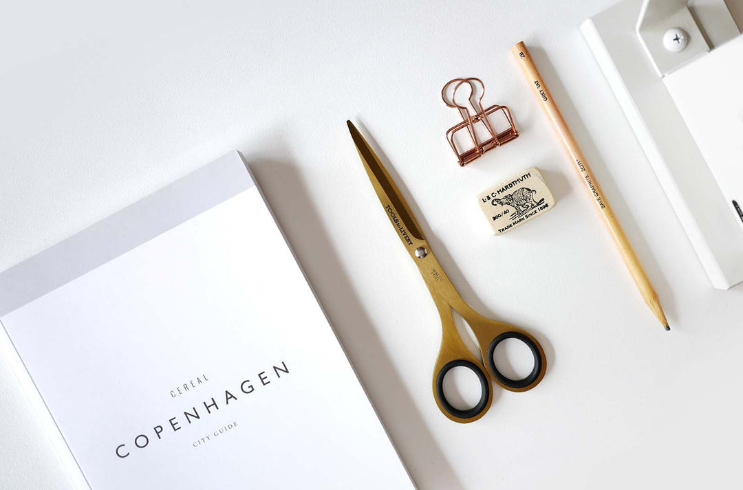 Premium overhead flat lay featuring the TOOLS to LIVEBY 6.5-inch gold scissors resting closed beside a Copenhagen travel guide, a pencil, and an eraser, styling a luxurious everyday carry workspace.