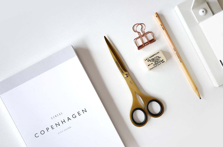 Premium overhead flat lay featuring the TOOLS to LIVEBY 6.5-inch gold scissors resting closed beside a Copenhagen travel guide, a pencil, and an eraser, styling a luxurious everyday carry workspace.