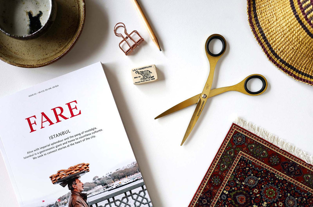 Travel-inspired flat lay showcasing the TOOLS to LIVEBY 6.5-inch gold scissors resting open next to a Fare magazine and everyday carry gear, embodying a luxurious mobile journaling setup.