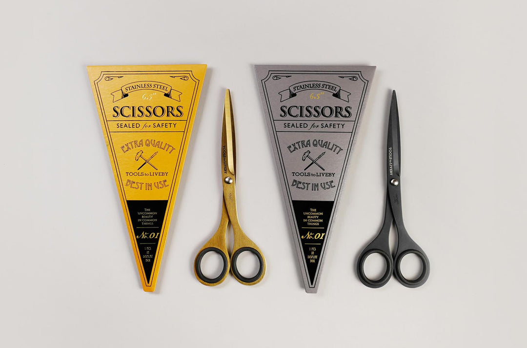 A collection of TOOLS to LIVEBY compact 6.5-inch scissors featuring striking gold and matte black finishes, displayed alongside their archival-style, vintage foil-stamped triangular cardboard sleeves in yellow and silver.