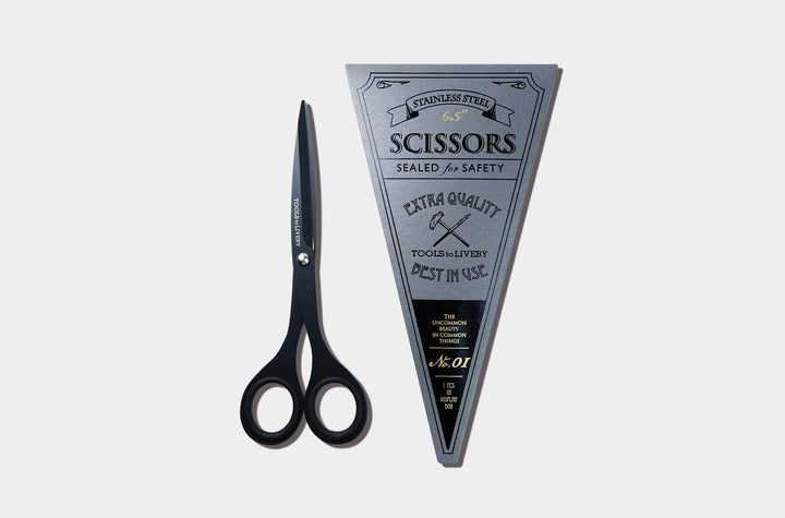 Overhead view of the compact TOOLS to LIVEBY 6.5-inch matte black scissors placed vertically next to its premium No. 01 silver triangular cardboard packaging sleeve with vintage letterpress details.