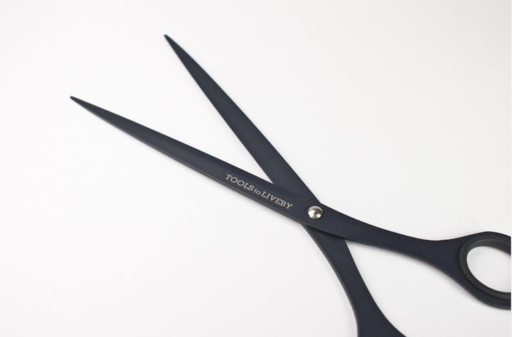 Macro detail of the partially open TOOLS to LIVEBY 6.5-inch matte black scissors, highlighting the engraved brand logo, the 'Stainless Steel' stamp on the inner blade, and the tactical ultra-matte finish.
