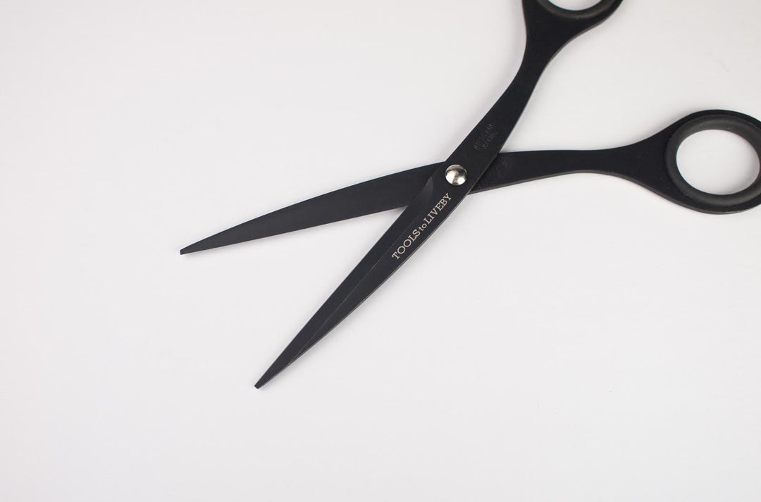 Macro close-up of the open TOOLS to LIVEBY 6.5-inch matte black scissors, displaying the sharp inner edge of the blades, the silver tension screw, and the precise anti-adhesive Teflon coating.