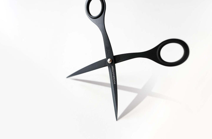 The TOOLS to LIVEBY 6.5-inch matte black scissors standing upright on their tips against a white surface, casting a sharp shadow that highlights the crisp lines of the Japanese stainless steel chassis.