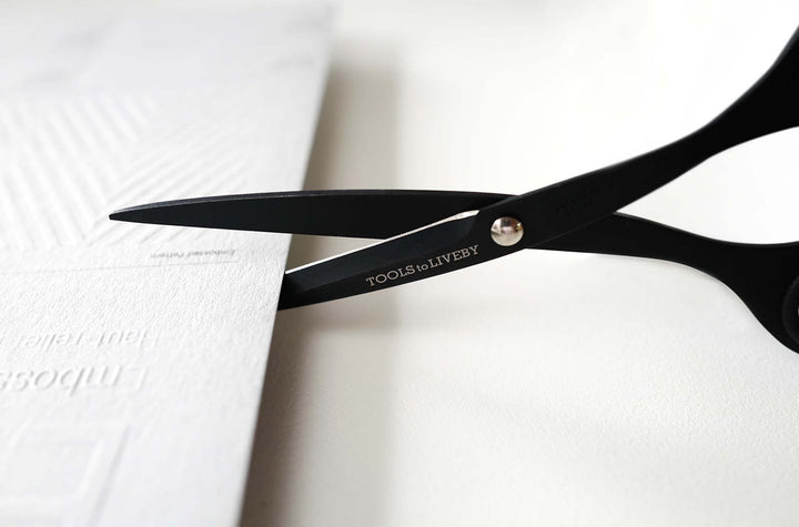 Close-up shot of the TOOLS to LIVEBY 6.5-inch matte black scissors effortlessly slicing through a thick sheet of embossed white paper, demonstrating the razor-sharp Japanese steel blades in action.