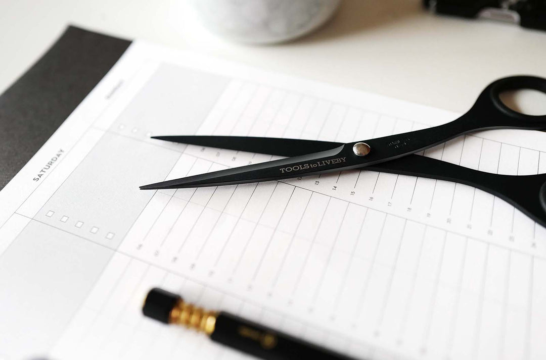 TOOLS to LIVEBY 6.5-inch matte black scissors resting open across a minimalist weekly planner, styled as an essential, pouch-ready everyday carry accessory for planning and journaling.