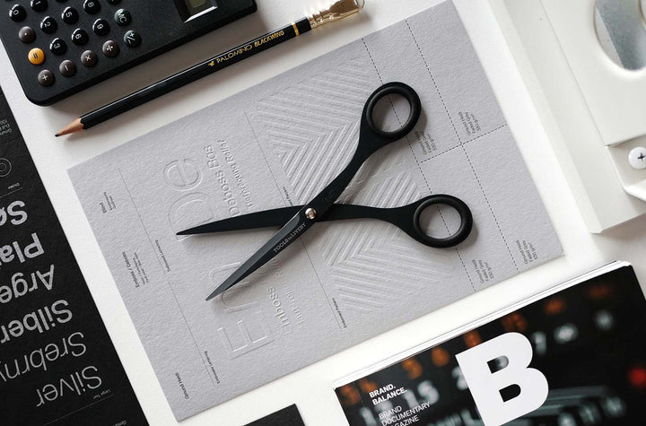 Premium overhead flat lay featuring the TOOLS to LIVEBY 6.5-inch matte black scissors resting open on textured grey paper, surrounded by a black pencil, calculator, and design magazine in a curated workspace.