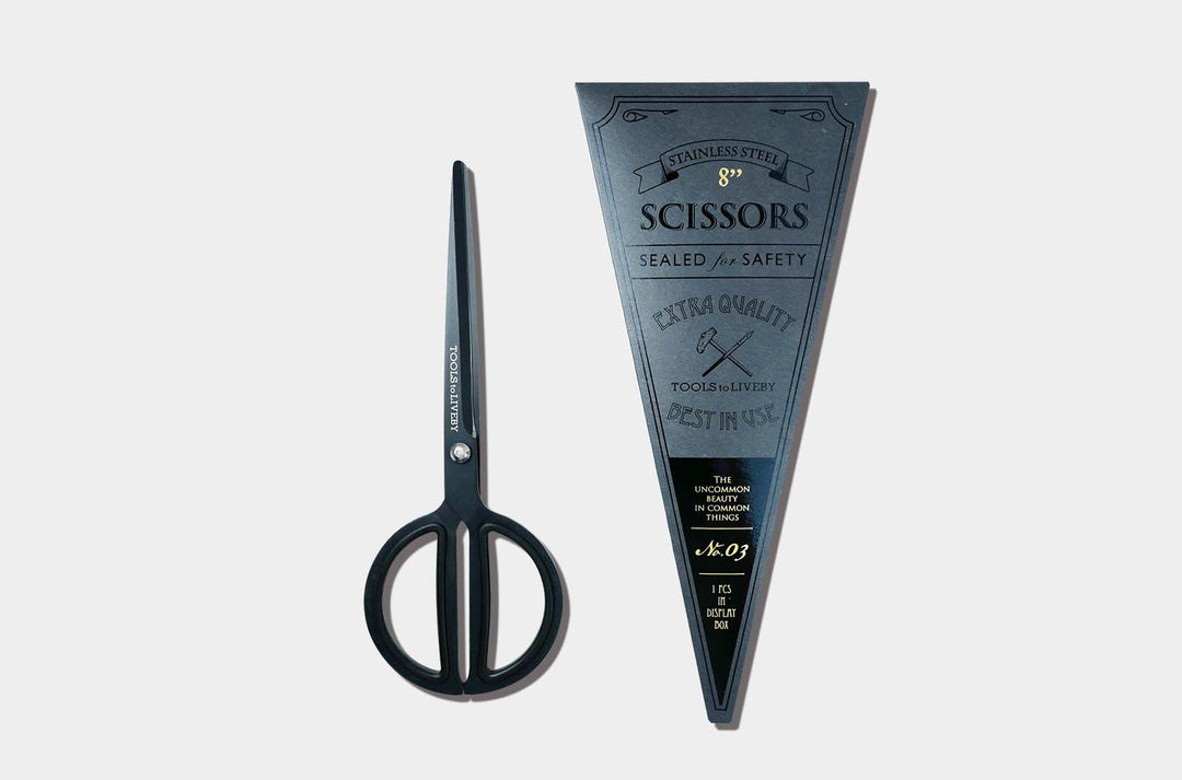 Overhead view of the TOOLS to LIVEBY 8-inch matte black scissors placed vertically next to its premium dark vintage foil-stamped triangular packaging sleeve.