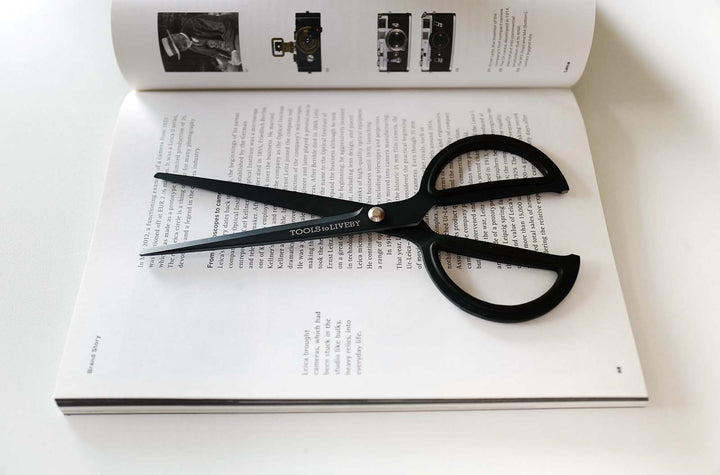 TOOLS to LIVEBY matte black scissors resting partially open across a photography book, styled as an essential, heavy-duty desktop accessory for creative professionals and studios.