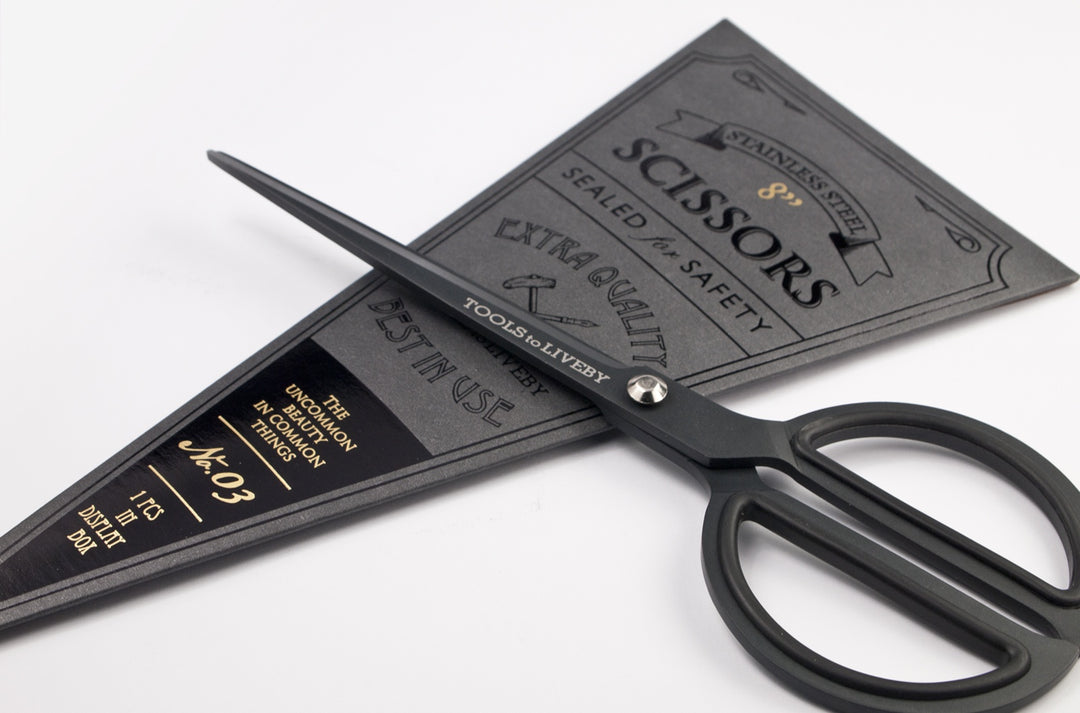 Close-up angled shot of the TOOLS to LIVEBY matte black scissors resting across its vintage dark cardboard sleeve, emphasizing the silver tension screw and engraved brand logo on the blade.