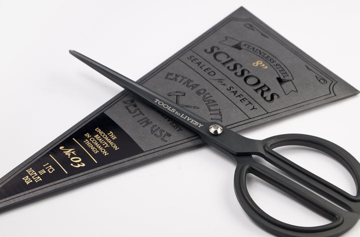 Close-up angled shot of the TOOLS to LIVEBY matte black scissors resting across its vintage dark cardboard sleeve, emphasizing the silver tension screw and engraved brand logo on the blade.