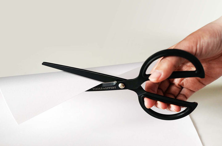 Close-up of a hand effortlessly slicing through crisp white paper using the TOOLS to LIVEBY 8-inch matte black scissors, demonstrating the razor-sharp Japanese steel blades and smooth cutting action.