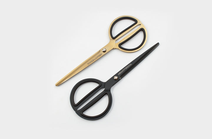 Two pairs of TOOLS to LIVEBY 8-inch premium desk scissors, one in stealthy ultra-matte black and one in gleaming gold, crossed diagonally on a clean white surface.