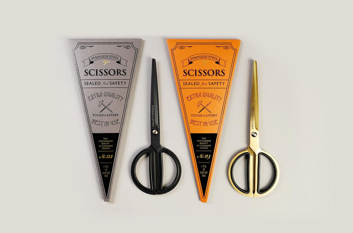 A collection of TOOLS to LIVEBY premium scissors featuring matte black and gold finishes, displayed alongside their archival-style, vintage foil-stamped triangular cardboard sleeves in silver and orange.