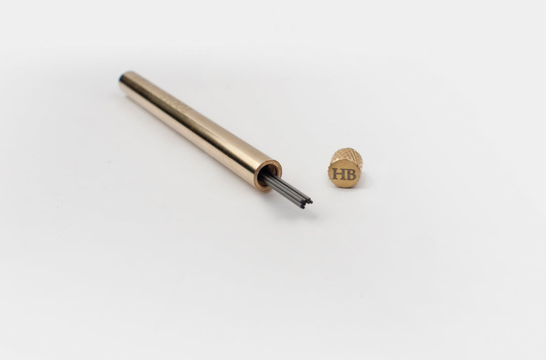 Uncapped TOOLS to LIVEBY solid brass lead holder displaying 0.5mm graphite refills inside, with the custom HB-grade knurled cap resting beside it on a clean white background.