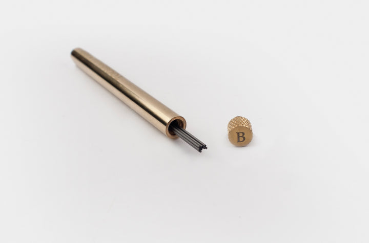 Uncapped TOOLS to LIVEBY solid brass lead holder displaying 0.5mm graphite refills inside, with the custom B-grade knurled cap resting beside it on a clean white background.