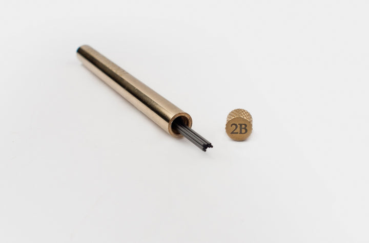 Uncapped TOOLS to LIVEBY solid brass lead holder displaying 0.5mm graphite refills inside, with the custom 2B-grade knurled cap resting beside it on a clean white background.