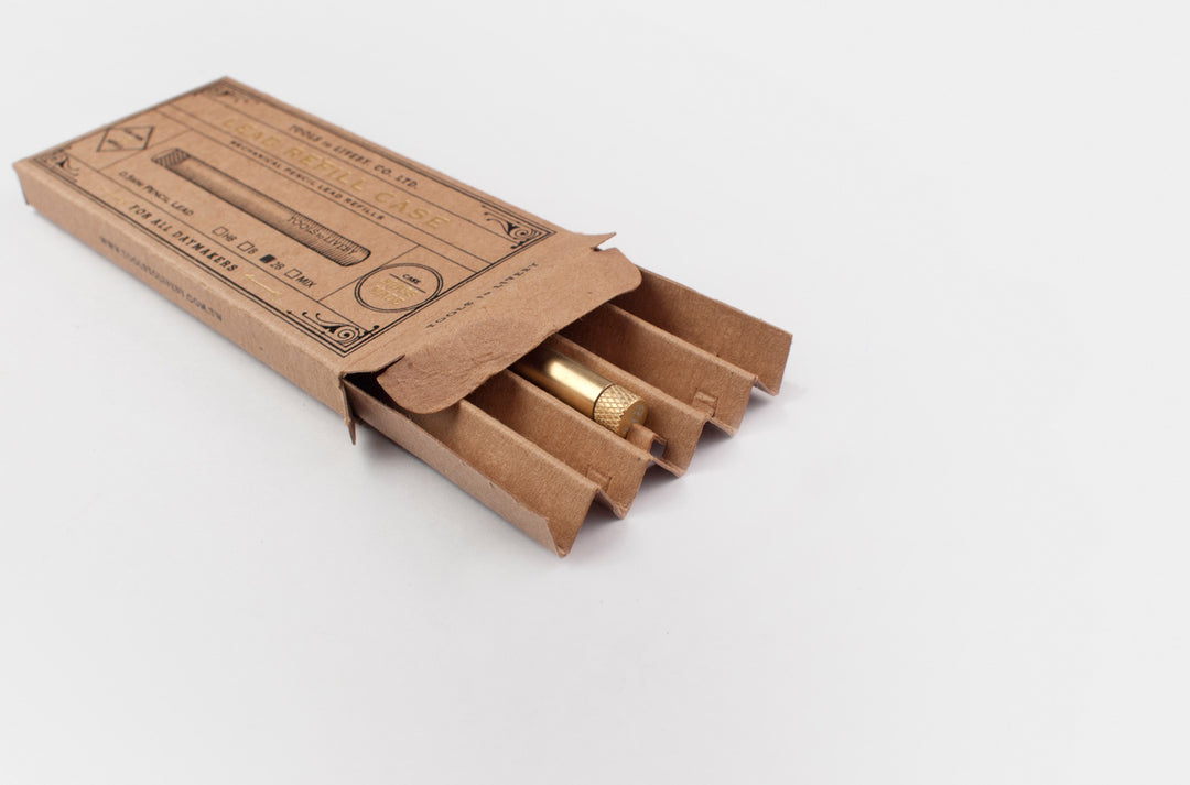 TOOLS to LIVEBY raw brass lead holder partially slid out of its vintage foil-stamped cardboard matchbox packaging on a minimalist white surface.