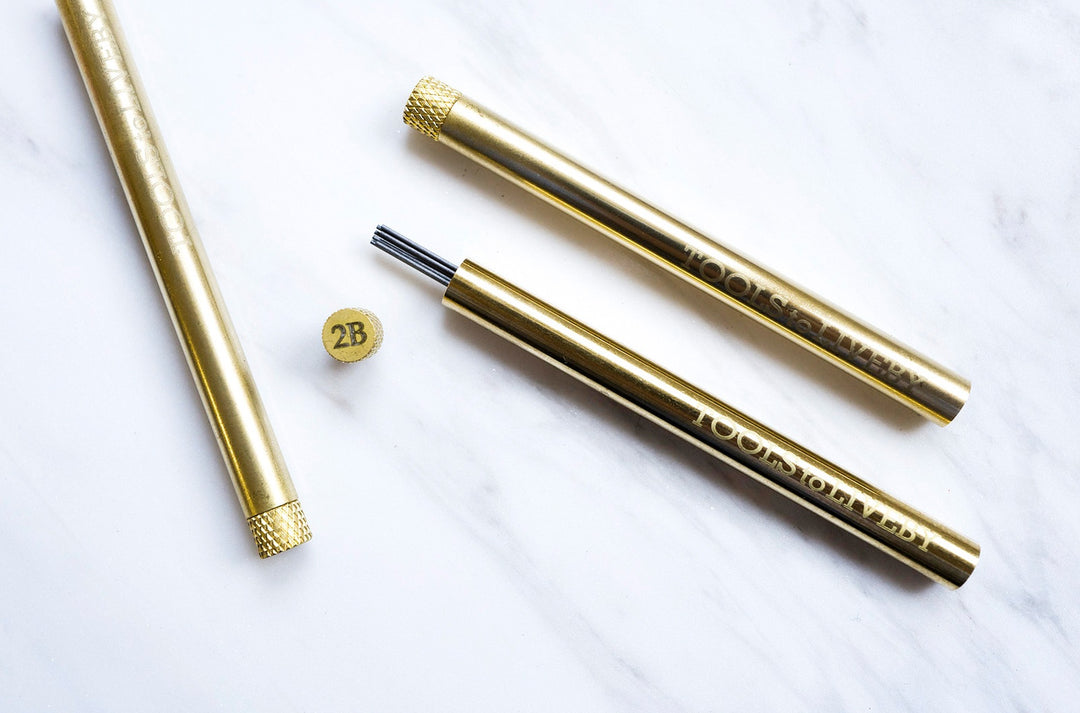Overhead view of two TOOLS to LIVEBY solid brass lead holders on white marble, showcasing the sleek cylindrical chassis, engraved brand logo, and a removed 2B grade cap exposing the protected graphite.