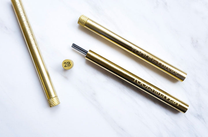 Overhead view of two TOOLS to LIVEBY solid brass lead holders on white marble, showcasing the sleek cylindrical chassis, engraved brand logo, and a removed 2B grade cap exposing the protected graphite.