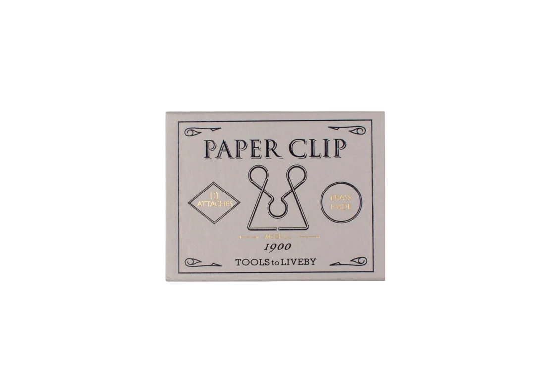 A top-down view of the closed, vintage-inspired light grey sliding matchbox for the TOOLS to LIVEBY McGill paper clip, showcasing elegant black typography with gold foil-stamped accents detailing its 1900 heritage design.